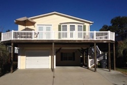 yellow/off-white 2 story house with large wrap-around deck on 2nd story, garage, and outdoor staircase; picture is taken from the paved driveway, pet friendly vacation rental in myrtle beach